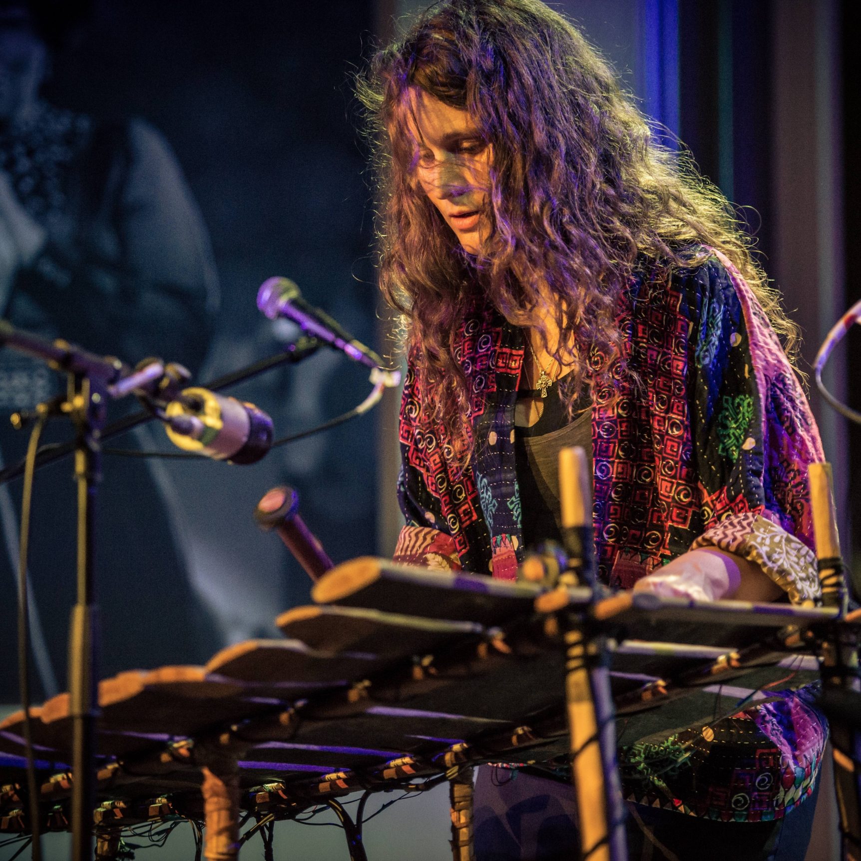 Bex Burch (young woman with brown, curly hair, colourful shirt) playing a large wooden instrument (the gyil) with beaters