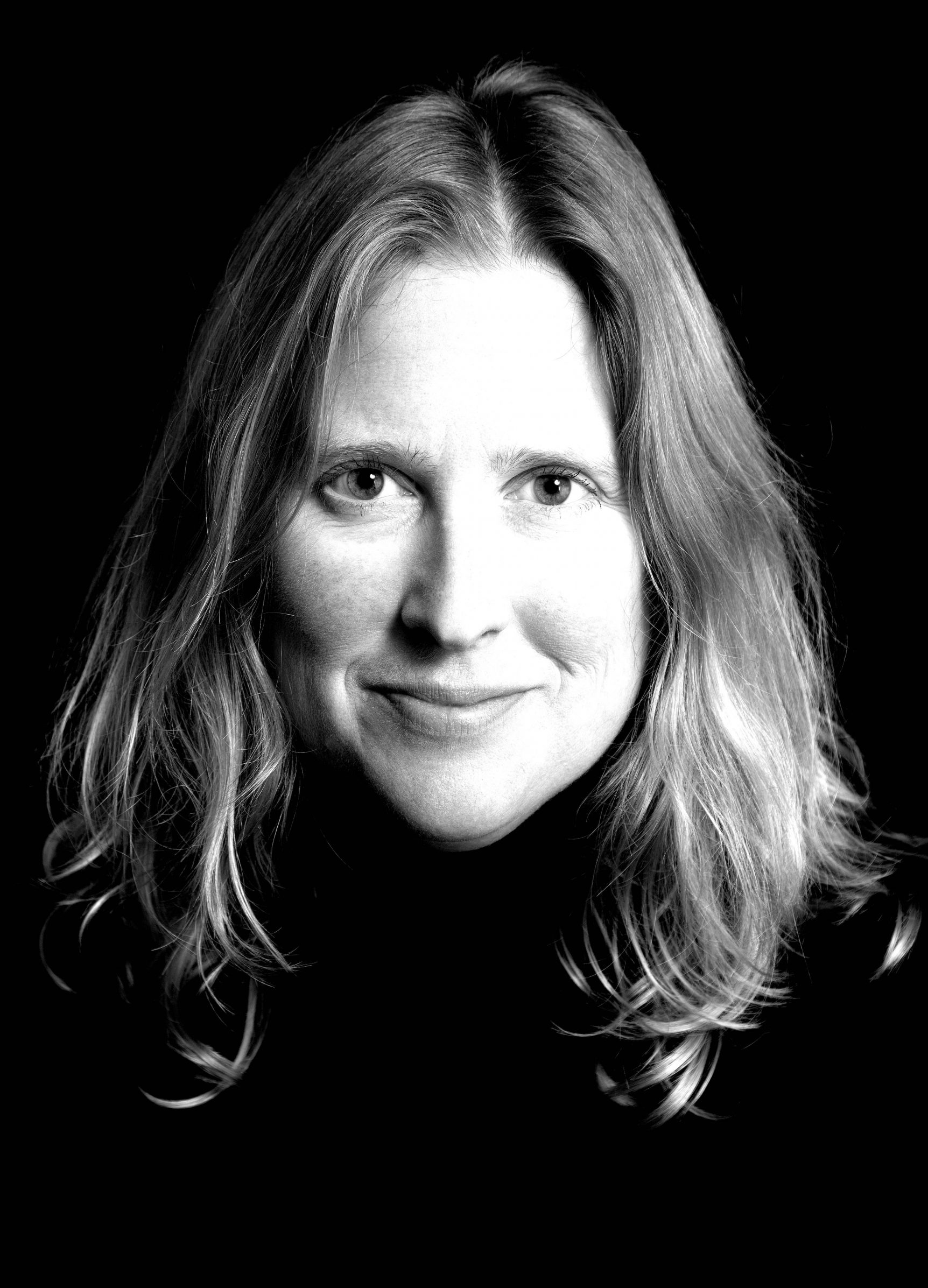 A close up black-and-white headshot with strong contrast. Charlotte smiles slightly at the camera, her black turtleneck blends with the black background while her light tone skin and hair pose a sharp contrast.