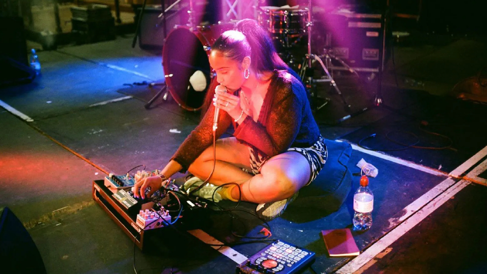 A young woman sits on stage using a microphone and adjusting an electronic instrument