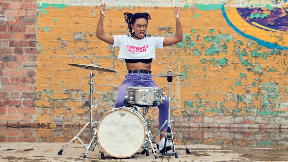 woman plays a drum kit and holds the drumsticks aloft