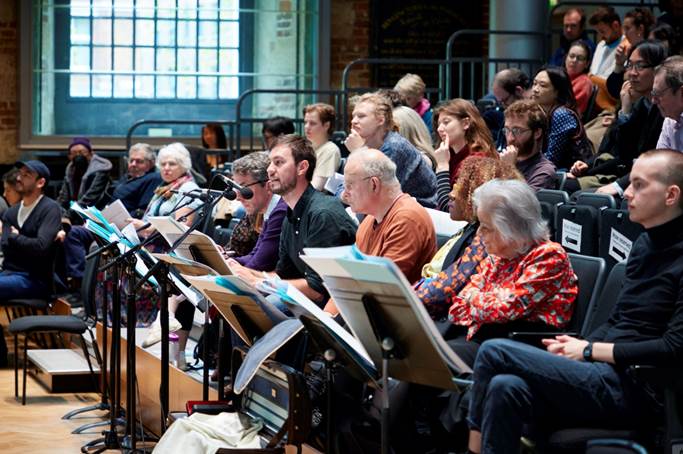 A photograph of an audience sat in an auditorium. There are mics and music stands, and the audience appear deep in thought.